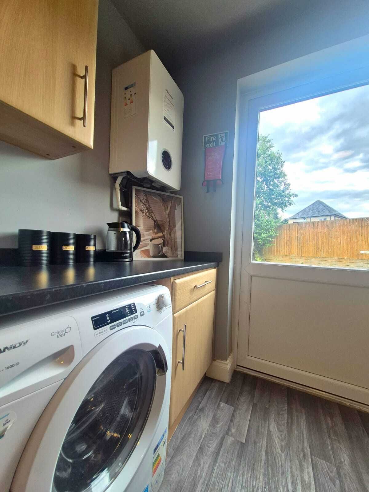 A compact kitchen with a washing machine, a coffee maker, and a window with a view of a backyard.