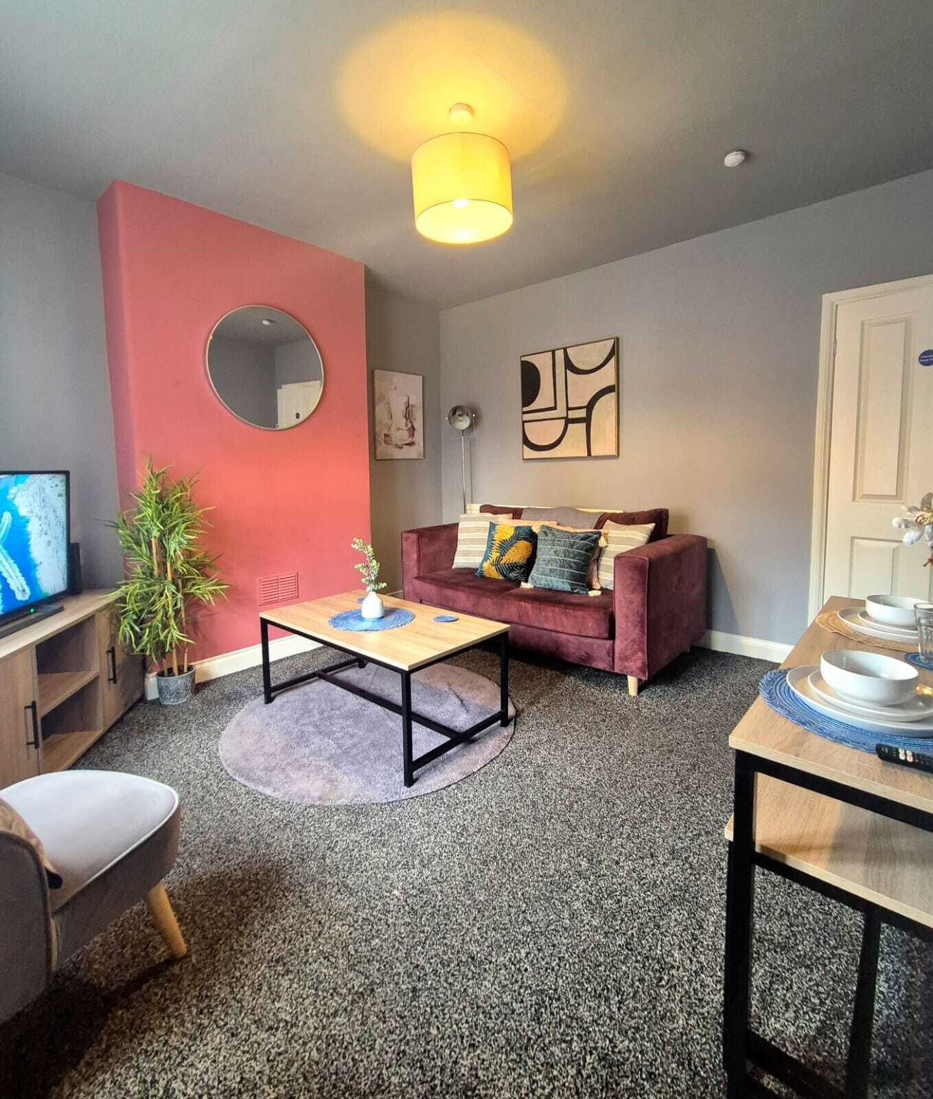 A living room with a red accent wall, a round mirror, a sofa with cushions, a coffee table, a TV, and a plant.
