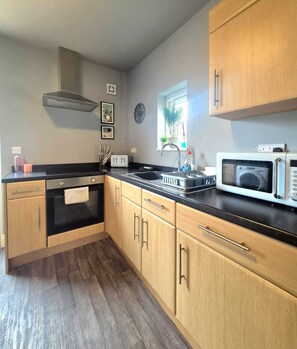 A kitchen with wooden cabinets, a black countertop, a microwave, and a window with a view of greenery. - Family 3BR Home | Free Parking | Near City Centre (Birstall)