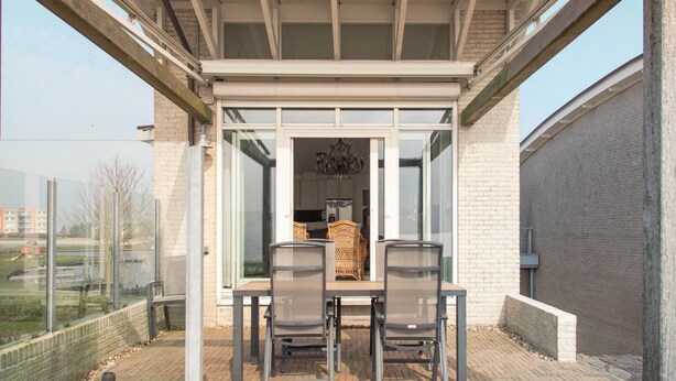 An outdoor patio with a table and chairs, a glass door leading to a room, and a view of a cityscape. - Panoramic Villa With Sauna & Lake Views (Makkum)
