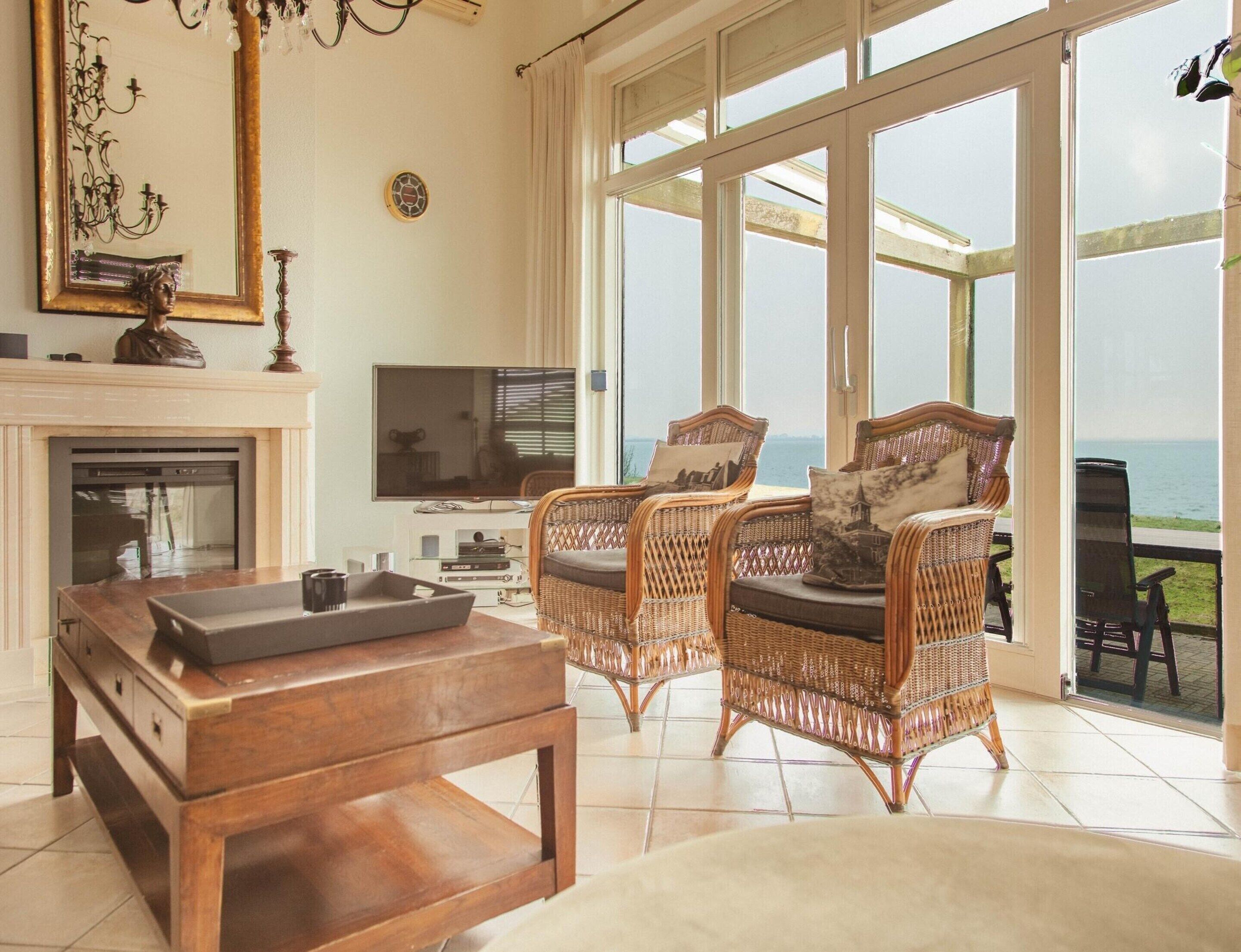 A spacious living room with a fireplace, a television, wicker chairs, and a large window overlooking the sea.