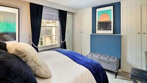 Ein Schlafzimmer mit einem Bett, einem fensterbelaubten Fenster, einem samtenen Ottomanen und einem Wandbild.