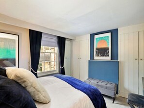 A bedroom with a bed, a window with curtains, a tufted ottoman, and a wall-mounted picture. - Luxury Stone Painswick Retreat for 6 (Stroud)