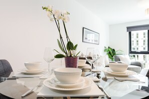 Ein Esstisch, gedeckt mit weißen Tellern, Schüsseln und Besteck, dazu eine Vase mit einer violetten Orchidee.