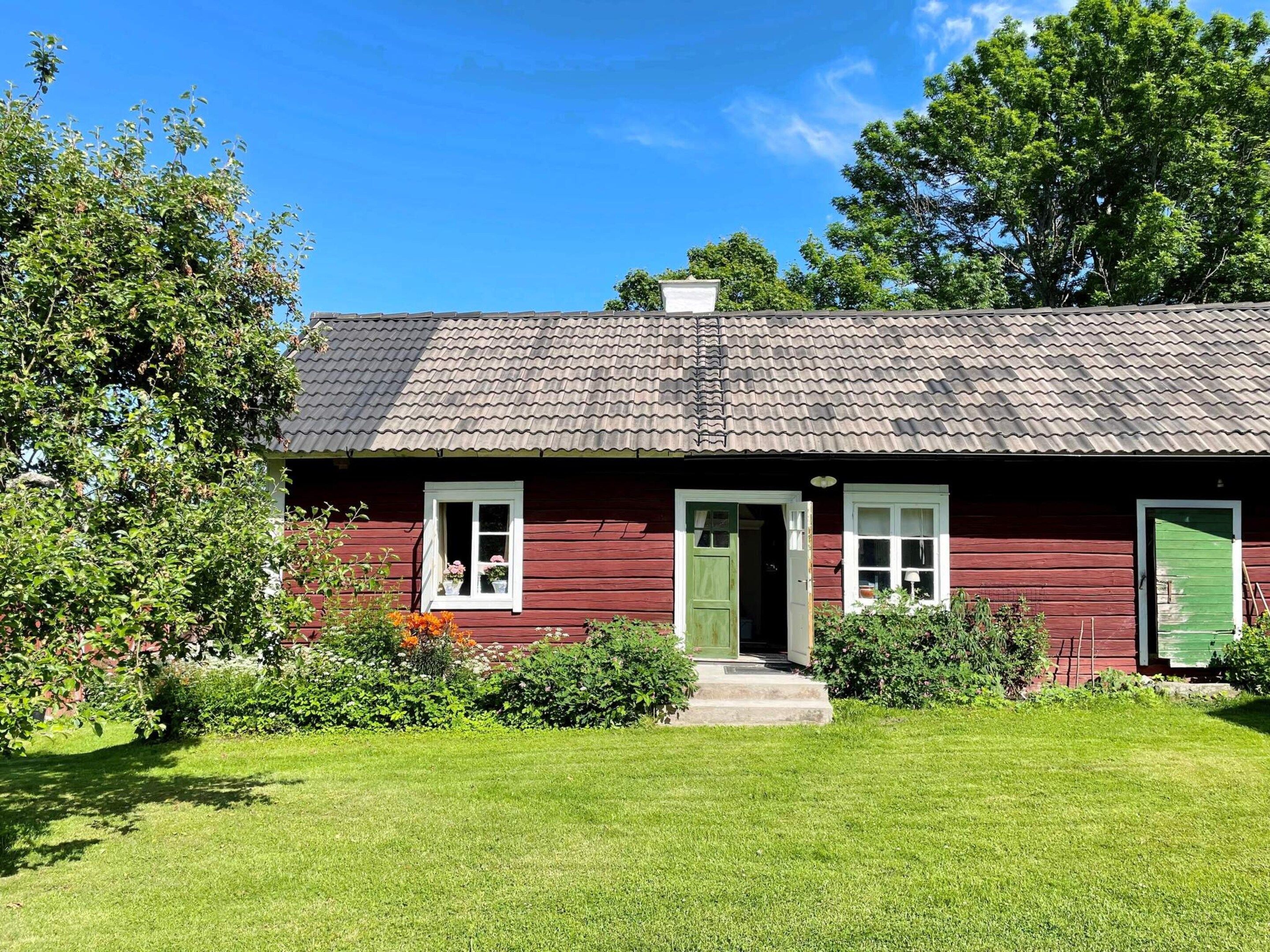 A red wooden house with a green door and white shutters, surrounded by a well-maintained lawn and trees.