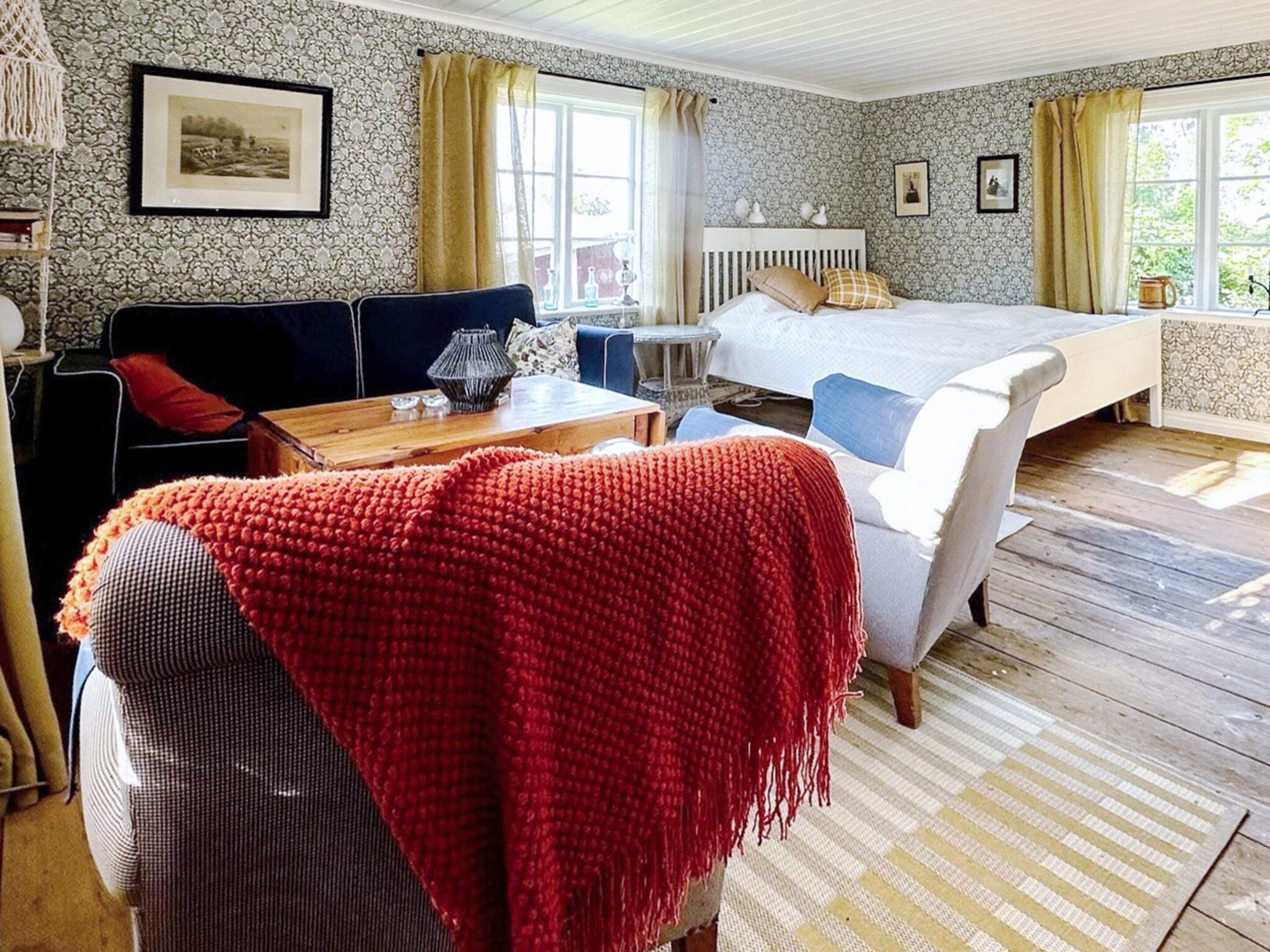 Ein gemütliches Zimmer mit Bett, einer Couch, einem Tisch und einem Sessel. Vorhänge, ein gerahmtes Bild an der Wand und ein Fenster mit Aussicht sind vorhanden.