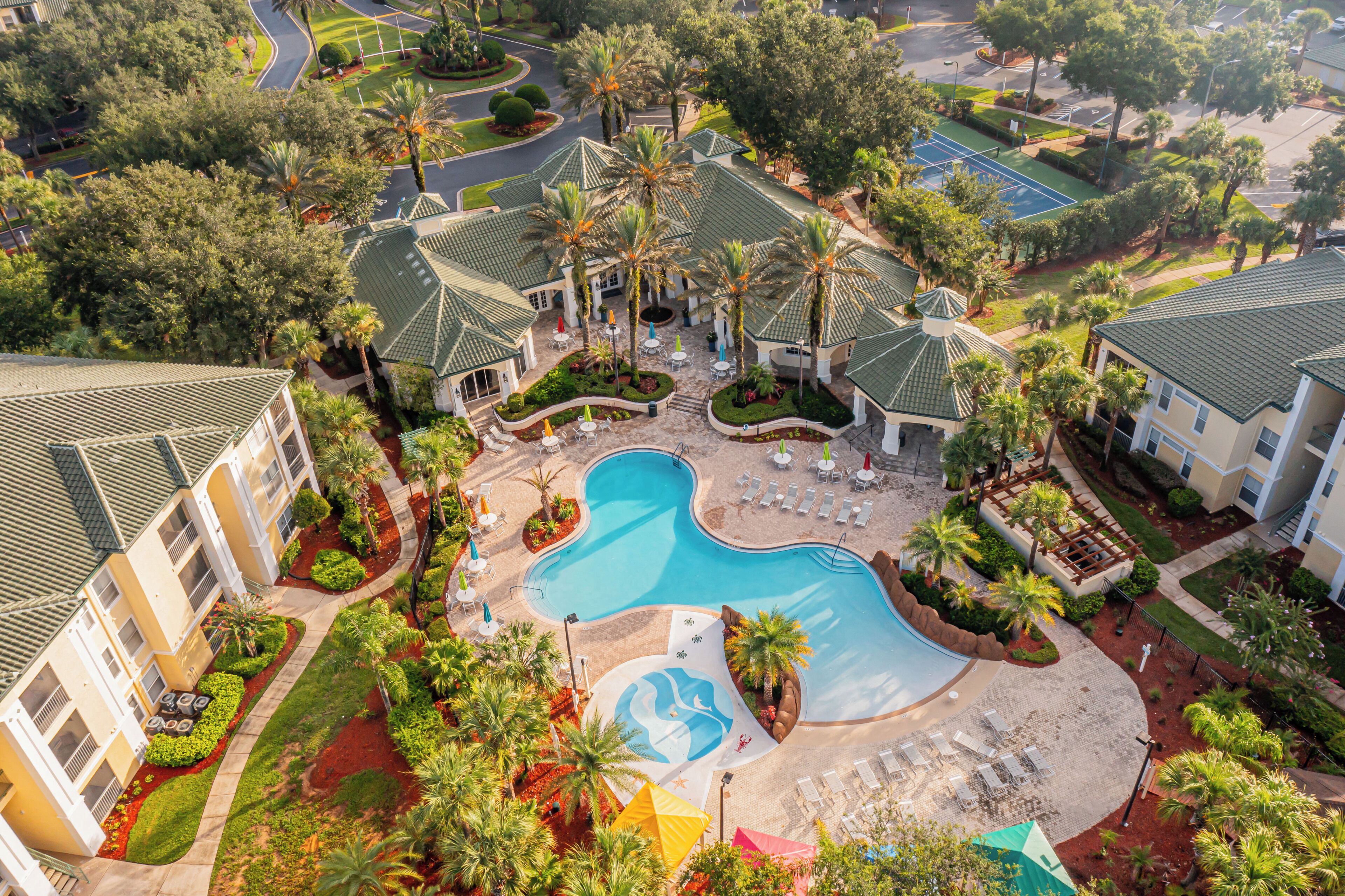 Vista aérea de un resort con piscina central, varias edificaciones de techos verdes y áreas ajardinadas circundantes.