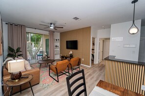 Uma sala de estar moderna com um sofá, cadeiras, uma mesa de centro e uma TV. Há uma varanda com cortinas e vista para o exterior.