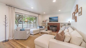 Un salon comprenant un canapé, un fauteuil et un pouf, une cheminée et une vue dégagée sur l’extérieur grâce à une grande fenêtre.