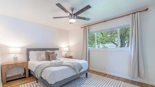 Une chambre à coucher avec un lit, des tables de chevet, un ventilateur de plafond, une fenêtre avec des rideaux et une vue sur les arbres à l’extérieur.