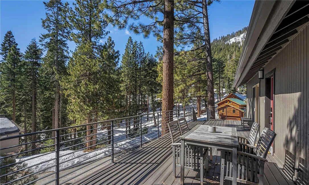 A wooden deck with a table and chairs, overlooking a forested area with tall trees and a cabin in the background.