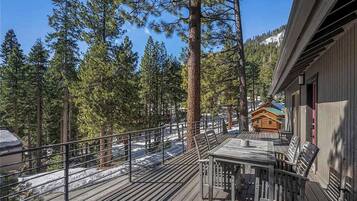 A wooden deck with a table and chairs, overlooking a forested area with tall trees and a cabin in the background.
