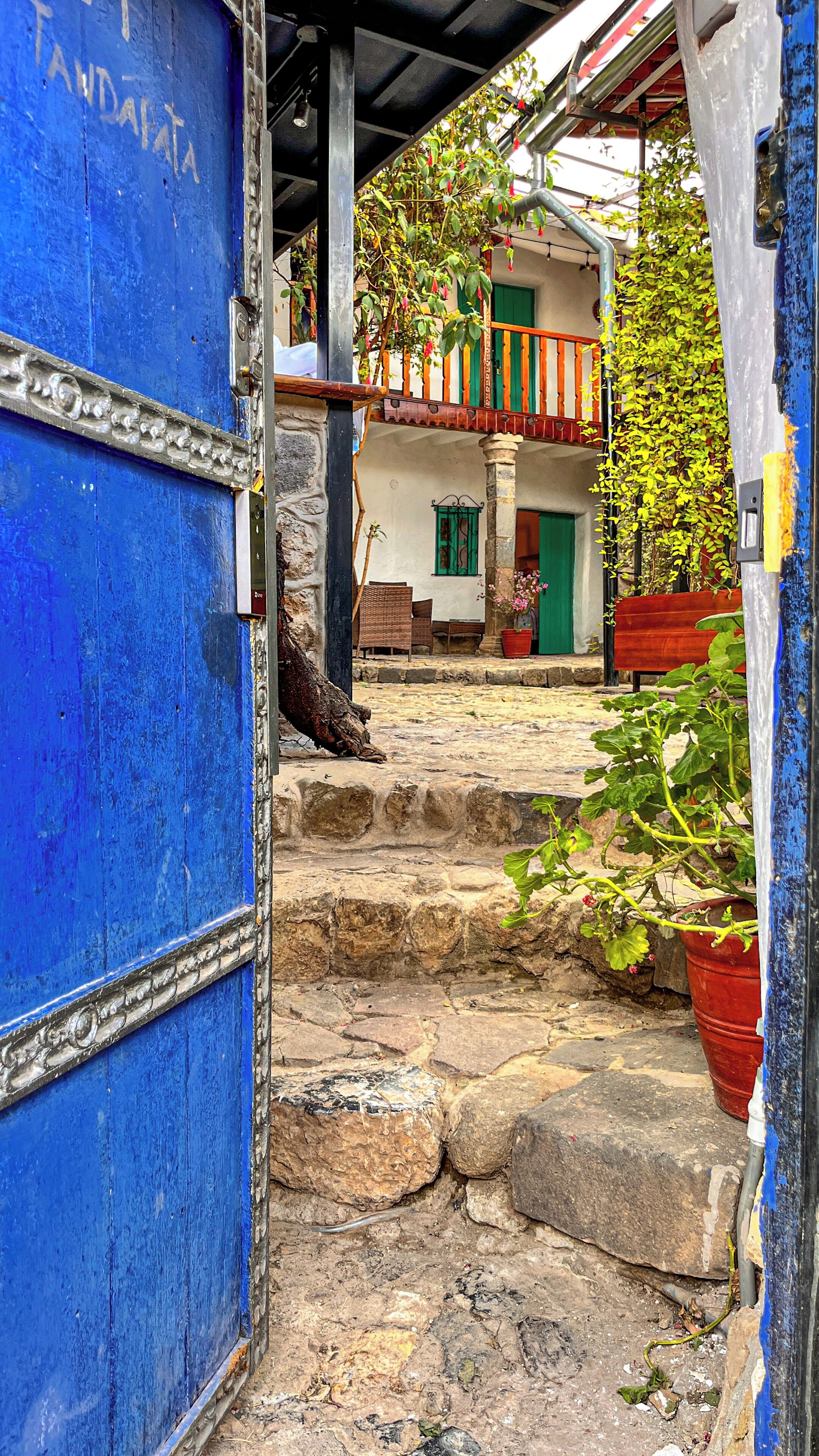 A stone staircase leading to a building with a blue door, a balcony with plants, and a red bench.