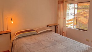 A room with a bed, a bedside table, a lamp, and a window with curtains.