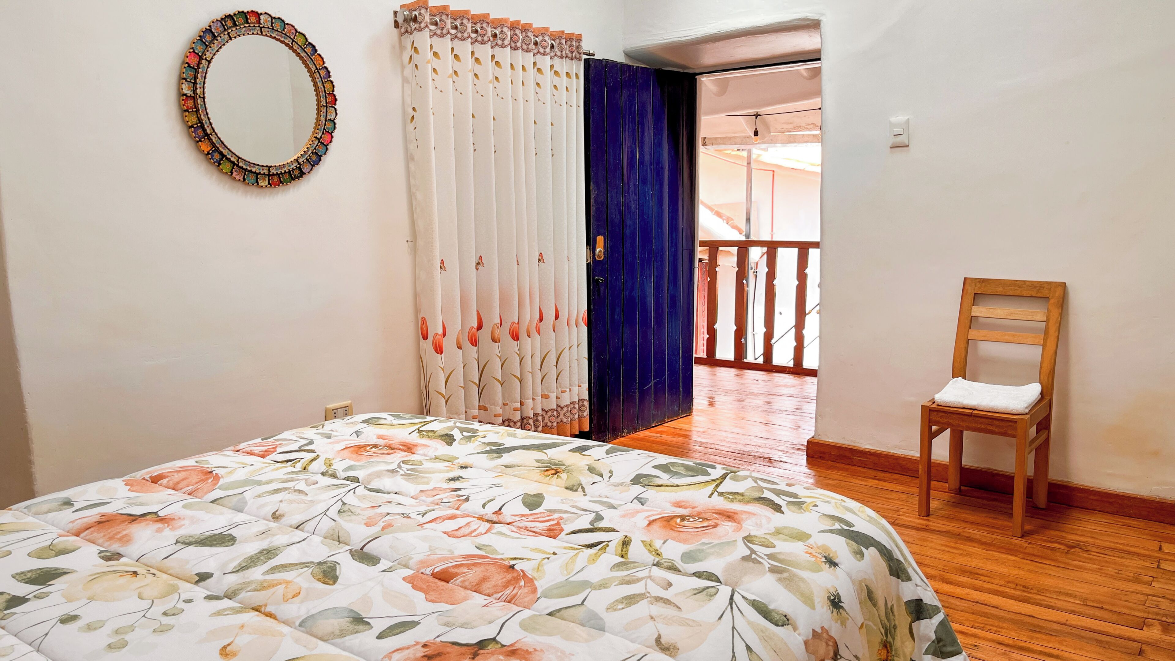 A bedroom with a floral bedspread, a wooden chair, a mirror, and a view of a balcony.
