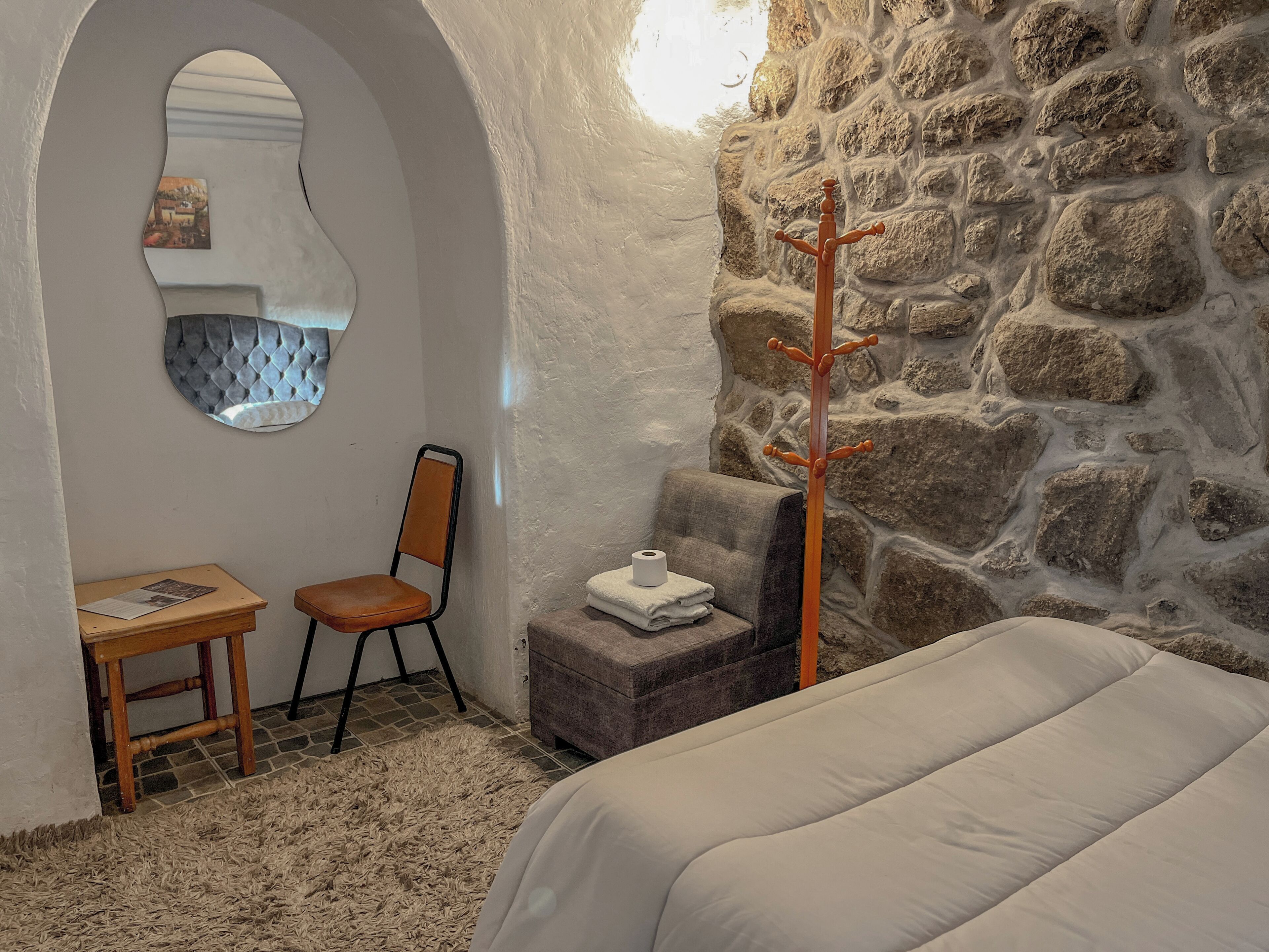 A cozy room with a stone wall, a wooden coat rack, a cushioned bench, a chair, and a small table with a magazine.