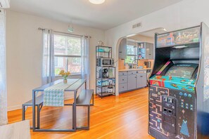 A kitchen with a retro arcade machine, a dining table with a bench, and a window with curtains. - Midtown Sanctuary w Pac-man! (Reno)