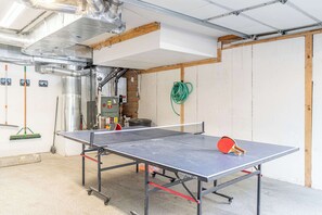 Uma mesa de pingue-pongue numa sala com tubulação aparente, uma vassoura e uma mangueira verde.