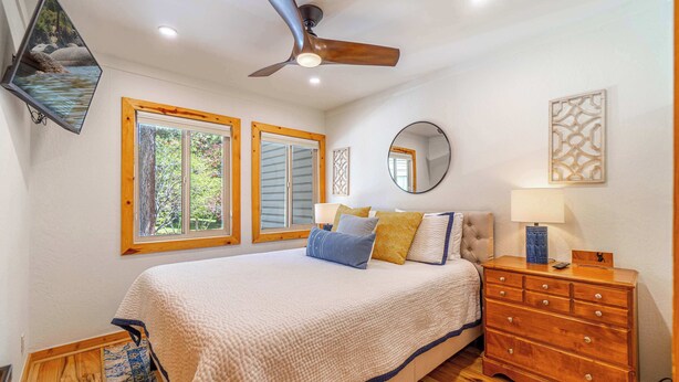 Quarto com cama, cómoda de madeira, ventoinha no teto e duas janelas com portadas.
