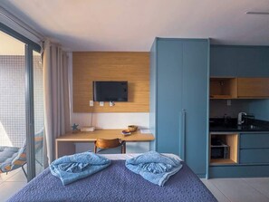 A modern hotel room with a bed, a desk, a television, and a kitchenette. - Próximo ao farol, 400m da praia, varanda & rooftop (Salvador)