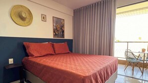 A hotel room with a large bed, a small table with chairs, a balcony with a view, and a wall with a hat and two pictures. - Studio c/ Varanda Vista Mar • Stella Maris & Farol (Salvador)