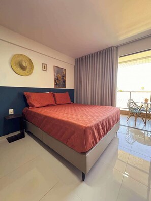 A hotel room with a large bed, a small table with chairs, a balcony with a view, and a wall with a hat and two pictures. - Studio c/ Varanda Vista Mar • Stella Maris & Farol (Salvador)