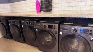 A row of black front-loading washing machines in a kitchen with white tiled walls and a granite countertop. - Studio c/ Varanda Vista Mar • Stella Maris & Farol (Salvador)