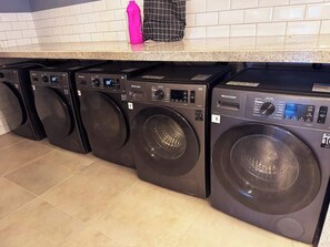 A row of black front-loading washing machines in a kitchen with white tiled walls and a granite countertop. - Studio c/ Varanda Vista Mar • Stella Maris & Farol (Salvador)