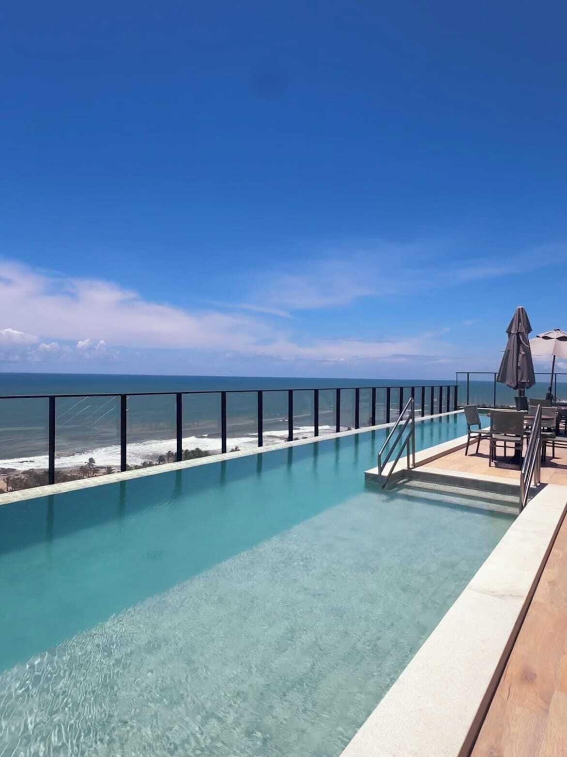 A clear blue pool with a view of the ocean, a wooden deck, and outdoor furniture.