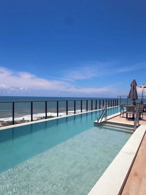 A clear blue pool with a view of the ocean, a wooden deck, and outdoor furniture. - Studio c/ Varanda Vista Mar • Stella Maris & Farol (Salvador)