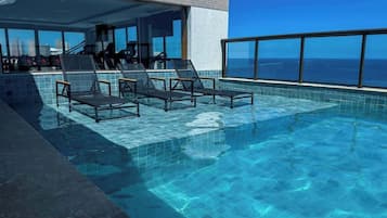 Una piscina infinita en la azotea con sillones de descanso, barandilla de cristal y vistas de la ciudad.