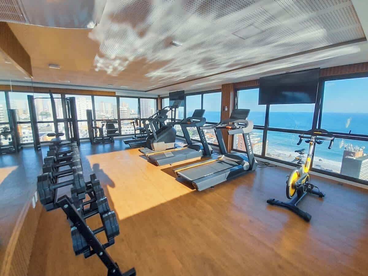 Un gimnasio con equipo de ejercicio y grandes ventanales con vistas a un paisaje urbano.