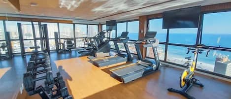 Un gimnasio con equipo de ejercicio y grandes ventanales con vistas a un paisaje urbano.