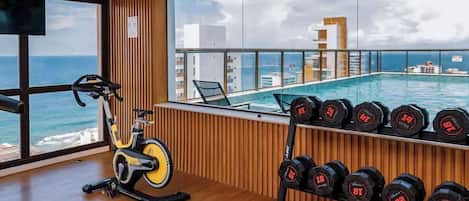 Un gimnasio con una bicicleta estática, una banca y un estante con mancuernas. Se ve una ventana grande con vistas al océano y edificios.