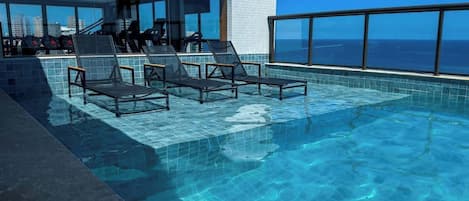 A rooftop infinity pool with lounge chairs, a glass balustrade, and a city view.