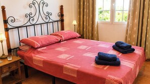 Ein ordentlich bezogenes Bett mit roter Bettdecke, zwei Kissen und gefalteten Handtüchern auf einem Nachttisch aus Holz, neben dem eine Lampe steht.