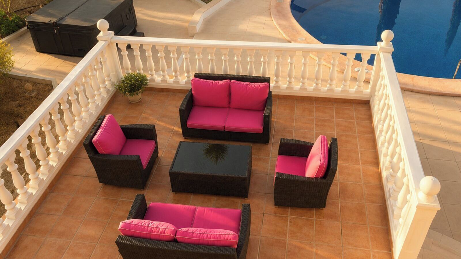 An outdoor patio with a pool, featuring a white balustrade, wicker furniture with pink cushions, and a potted plant.