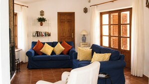 A living room with two blue sofas, a white armchair, a bookshelf, a wooden door, and a window with curtains.