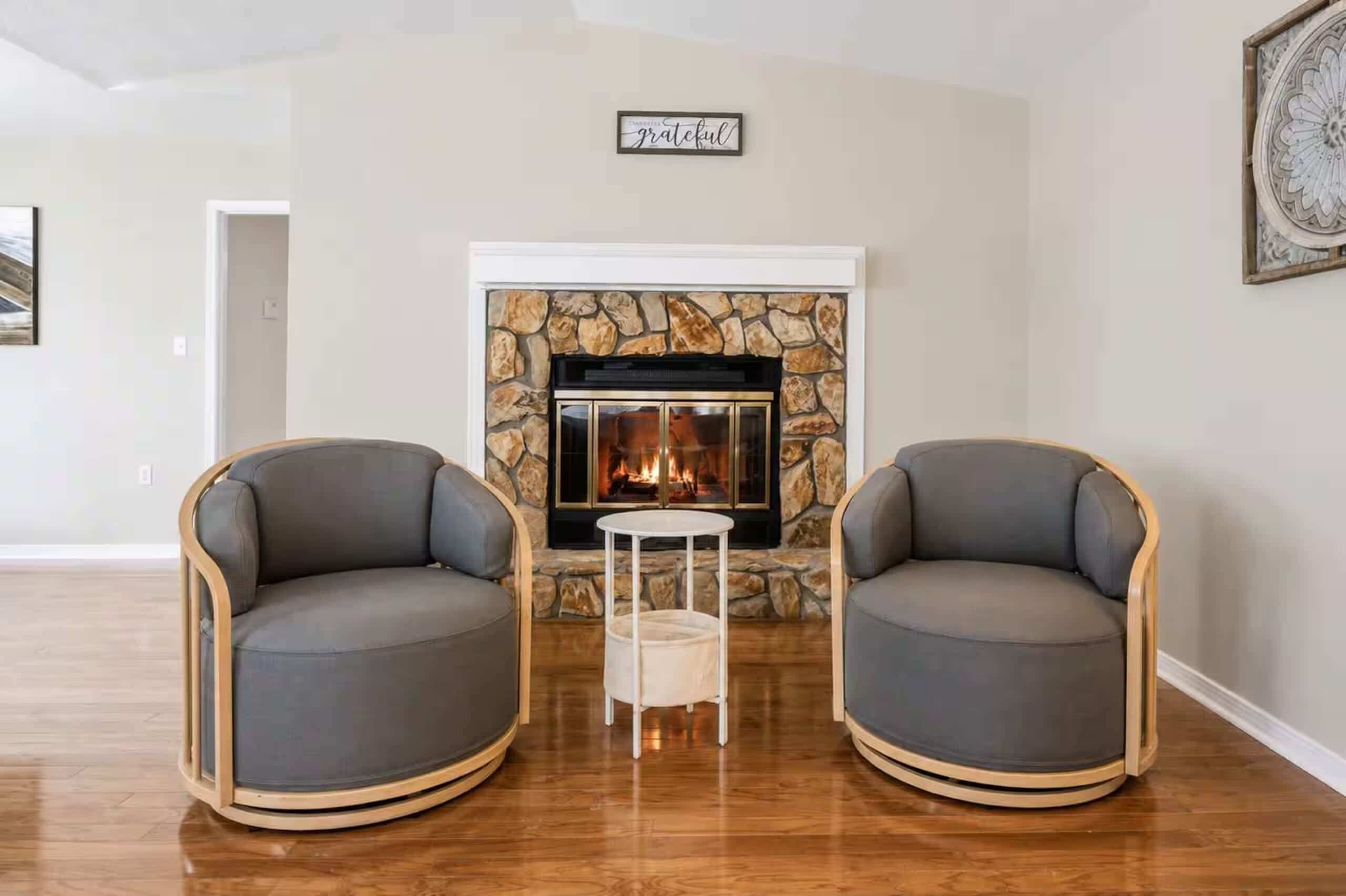 A room with a stone fireplace, two grey armchairs, and a small white table.