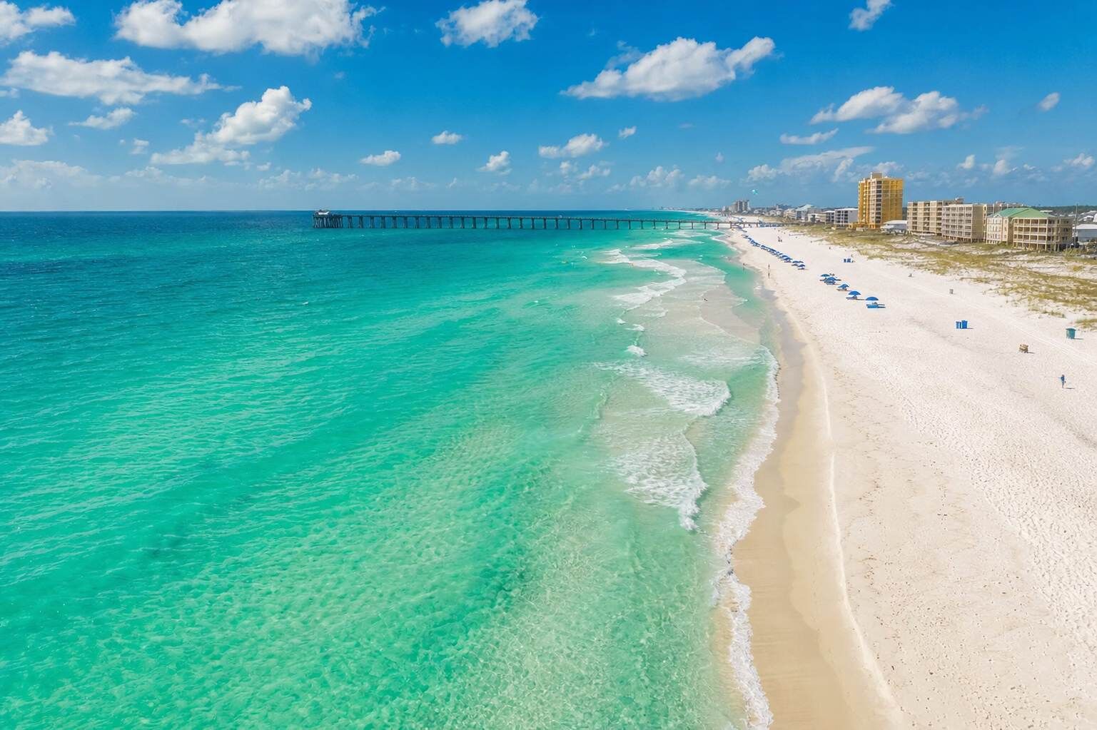 A beach with clear turquoise waters, a pier extending into the sea, and buildings along the shore.