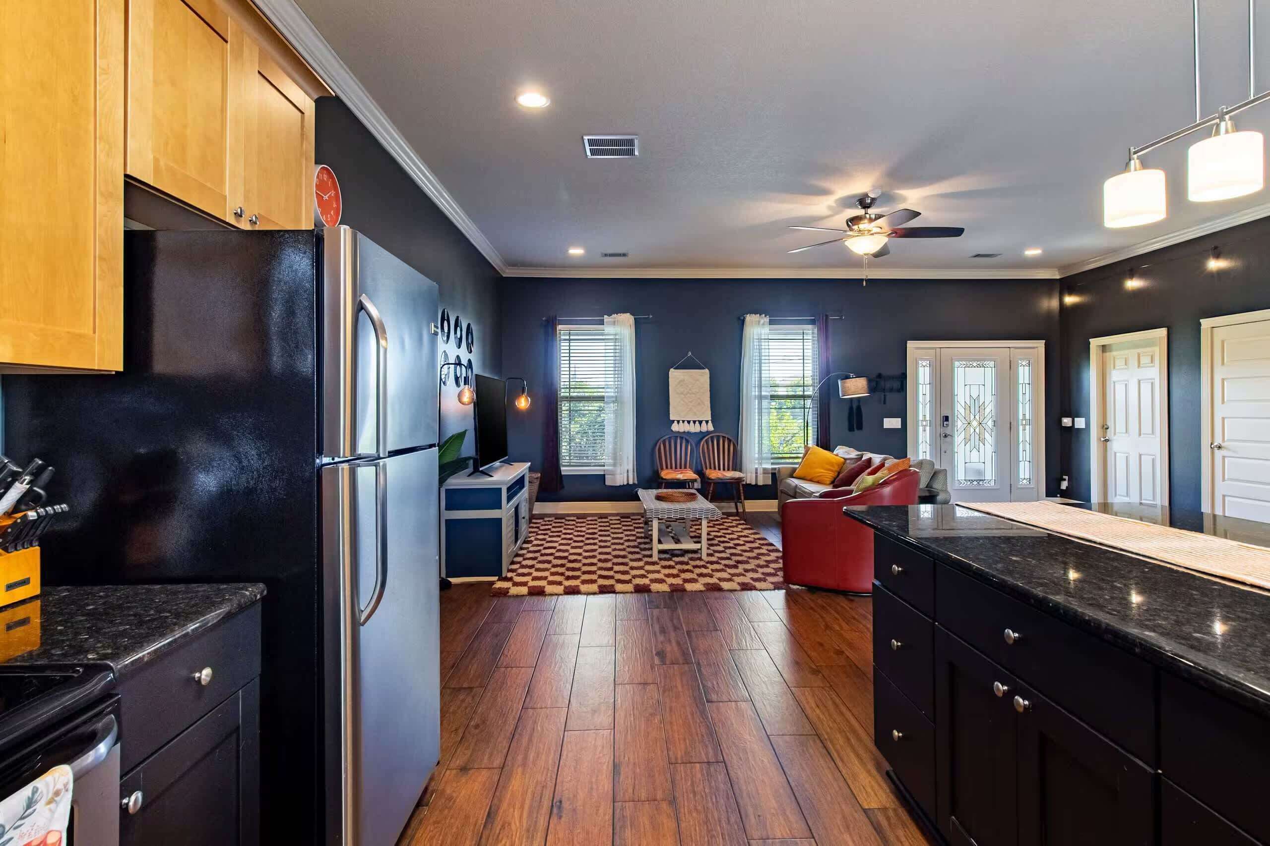 A modern kitchen with a refrigerator, stove, and wooden cabinets. There is a dining area with a table and chairs, and a living room with a sofa and a television.
