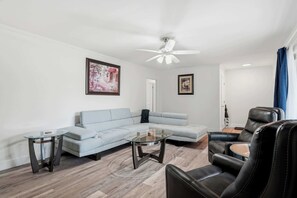 A modern living room with a light grey sofa, a glass coffee table, a black leather armchair, and a ceiling fan. - Indy 3BR | Game Room + Fire Pit | Sleeps 8 (Indianapolis)