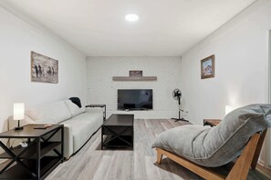 A modern living room with a white brick wall, a flat-screen TV, a sofa, a coffee table, a chair, and a side table with a lamp. - Indy 3BR | Game Room + Fire Pit | Sleeps 8 (Indianapolis)