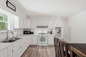 A kitchen with white cabinets, a stainless steel sink, a microwave, a stove, and a wooden dining table. - Indy 3BR | Game Room + Fire Pit | Sleeps 8 (Indianapolis)
