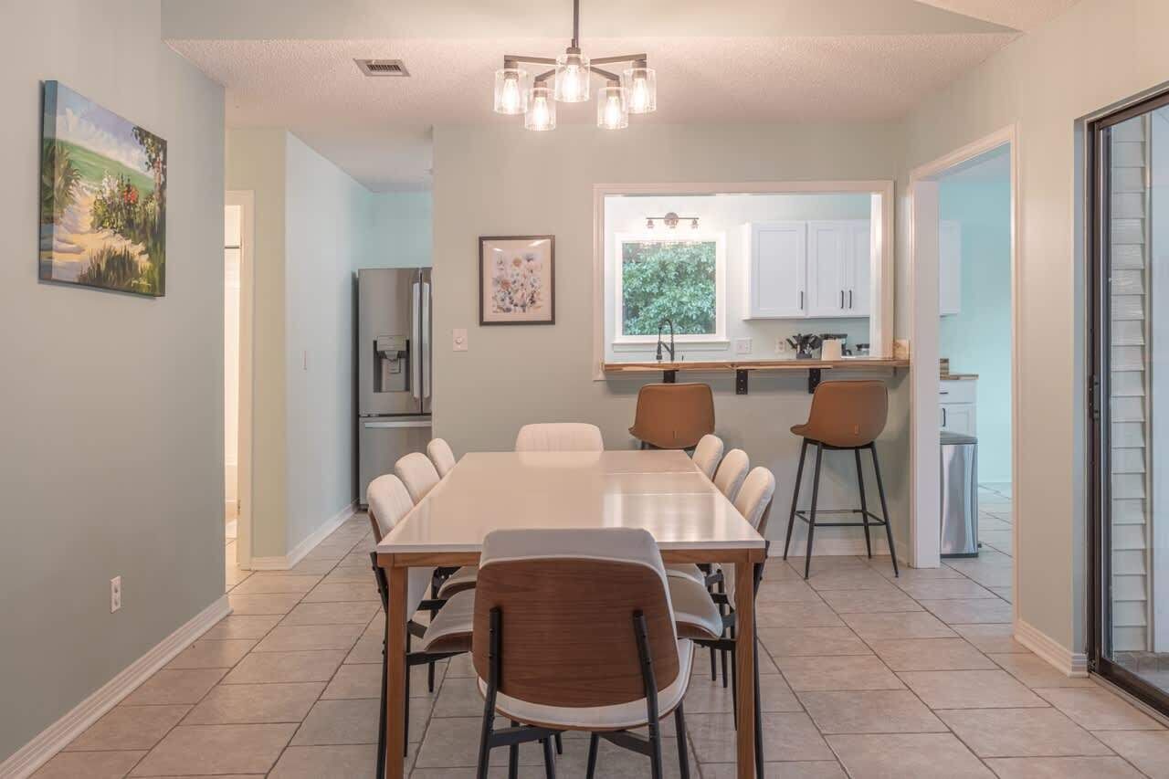 A dining area with a large table and chairs, a kitchen in the background, and a window with a view of greenery.