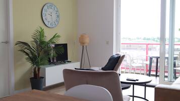 Un salon avec un canapé, une petite table, une plante en pot, un téléviseur et une horloge murale.