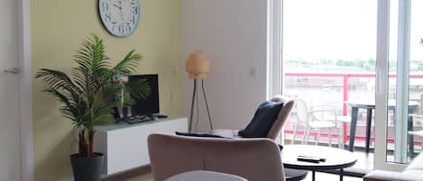 A living room with a sofa, a small table, a potted plant, a television, and a wall clock.