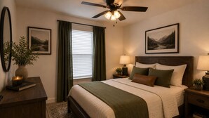 A bedroom with a bed, a ceiling fan, two bedside tables with lamps, a nightstand, a window with curtains, and two framed pictures on the wall. - The Washington Lofts (Ellicottville)