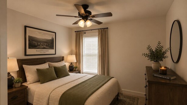 A neatly arranged bedroom with a bed, two bedside tables with lamps, a ceiling fan, a framed picture, a window with curtains, a bedside plant, a small cabinet with a book, and a mirror on the wall. - The Washington Lofts (Ellicottville)