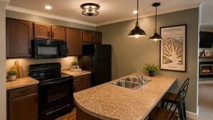 A modern kitchen with wooden cabinets, a black refrigerator, a built-in oven, a granite countertop, and pendant lighting. - The Washington Lofts (Ellicottville)